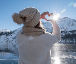 Frau in Winterkleidung formt ein Herz mit den Händen vor einer sonnigen verschneiten Berglandschaft.